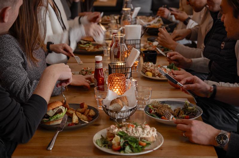 Gjester samlet rundt et langbord mens de nyter mat og drikke i en varm og sosial restaurantatmosfære.