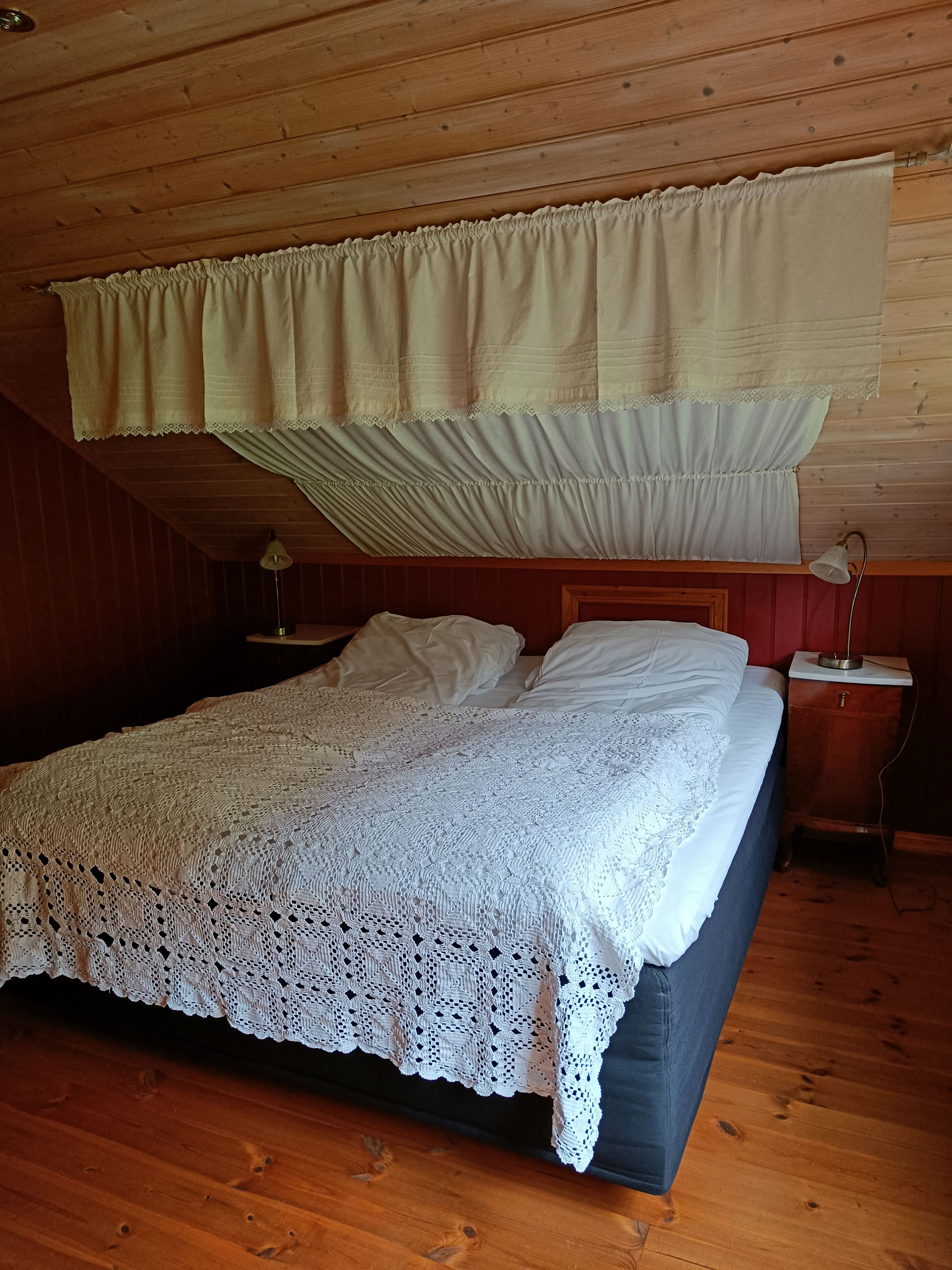 Orheimstunet in Nordfjord - Attic bedroom with a double bed, white lace bedspread, wooden ceiling and floor, and bedside lamps and tables..