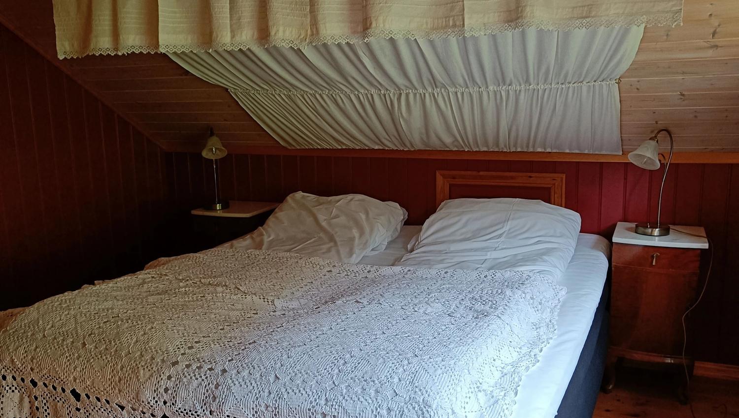 Orheimstunet in Nordfjord - Attic bedroom with a double bed, white lace bedspread, wooden ceiling and floor, and bedside lamps and tables..