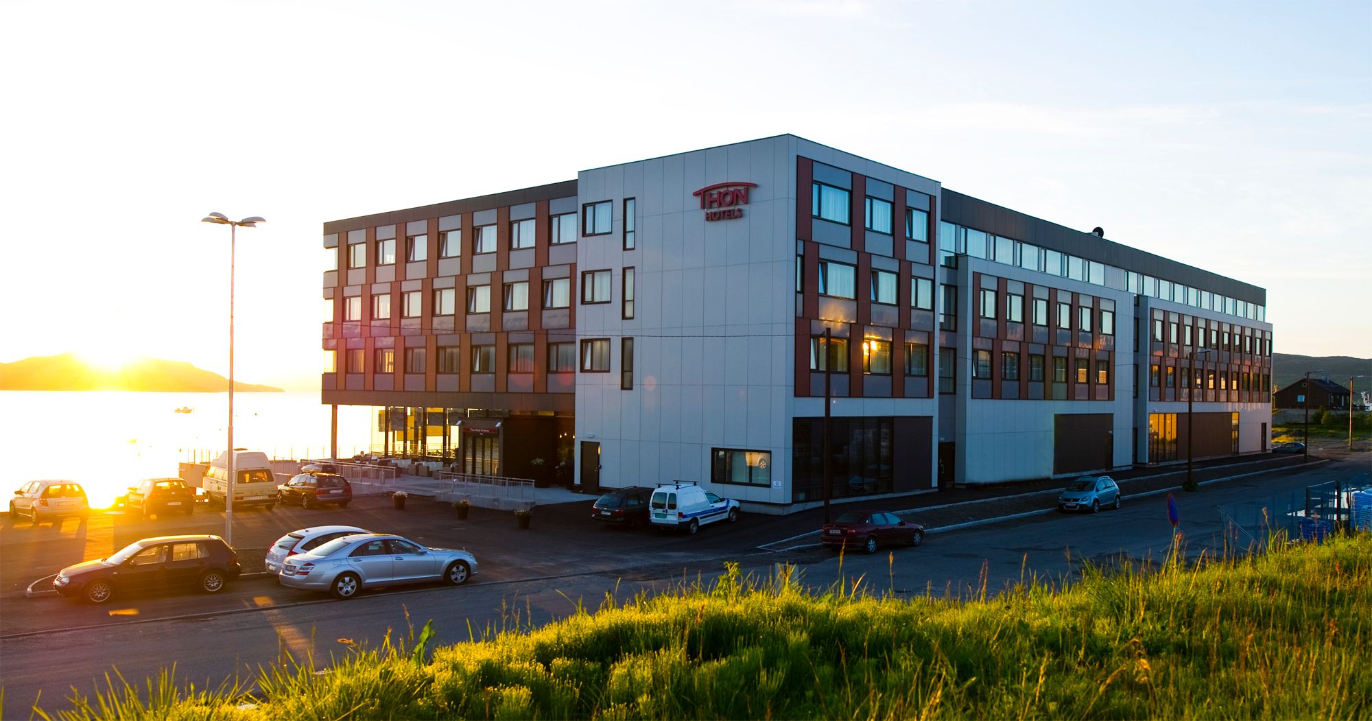 Modern hotel building at sunset with cars parked in front and a waterfront view.