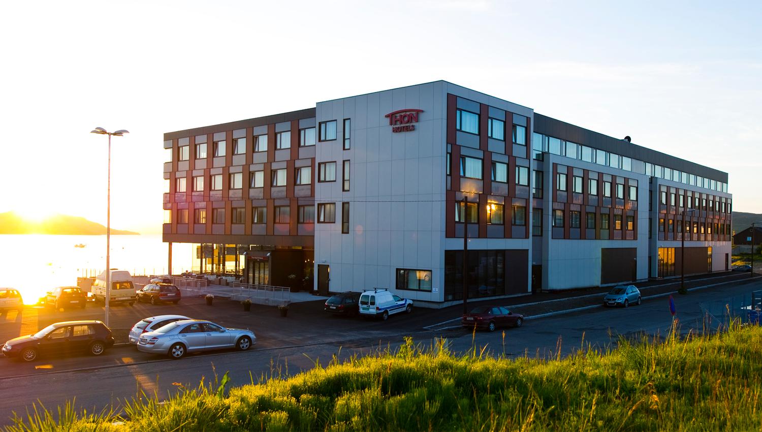 Modern hotel building at sunset with cars parked in front and a waterfront view.