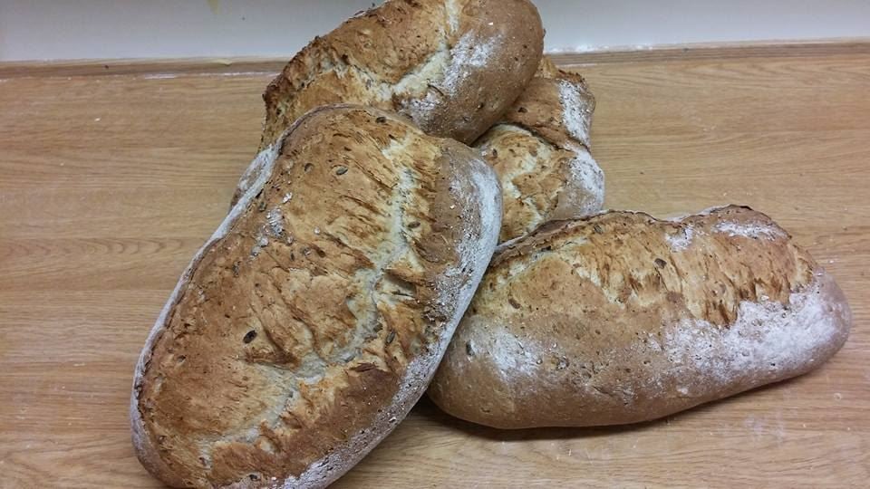 Several flour-dusted rustic bread loaves stacked on a wooden table, with a plain wall in the background.