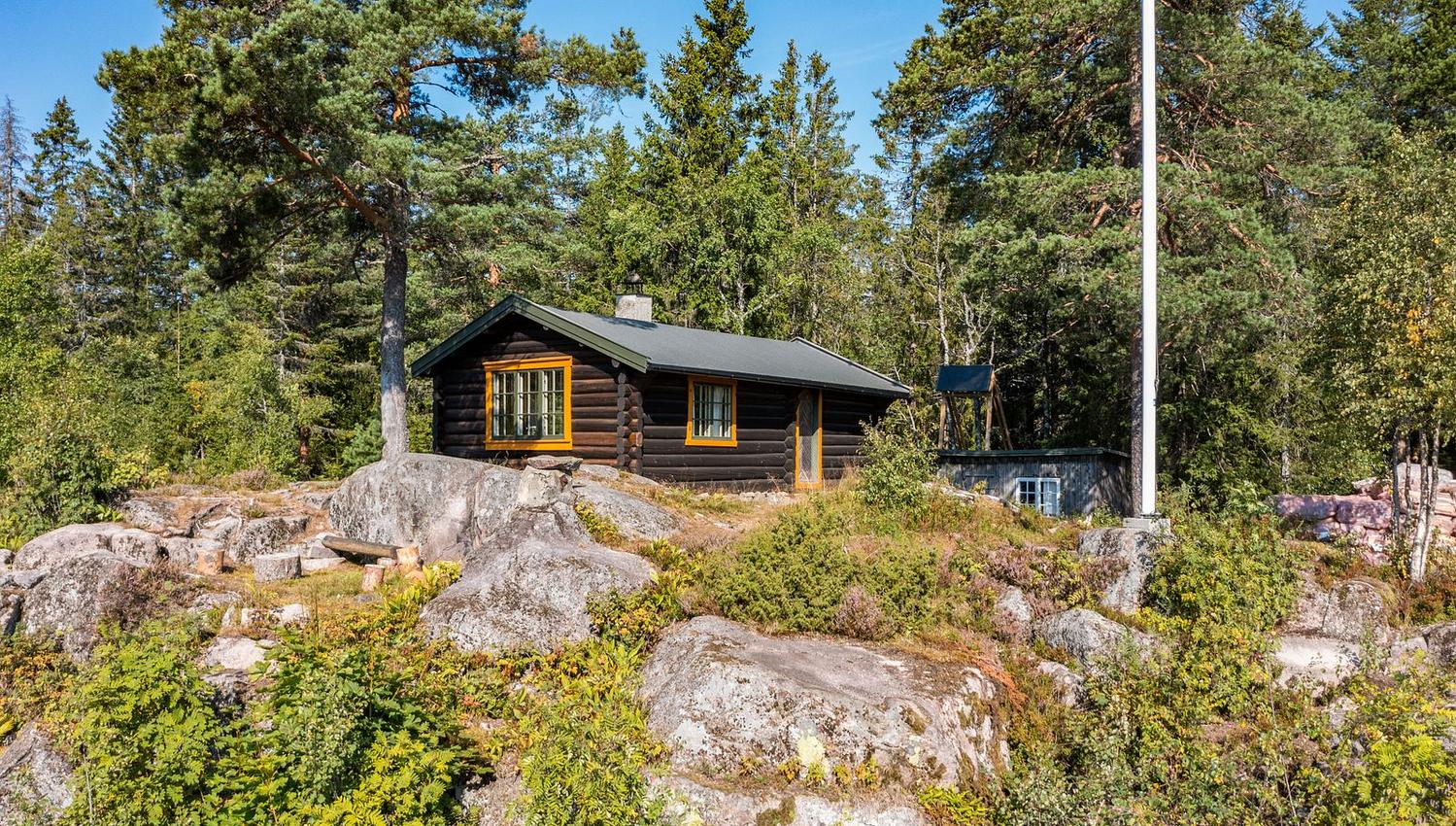 Brun markahytte og flaggstang på fjellknaus omgitt av grønn vegetasjon