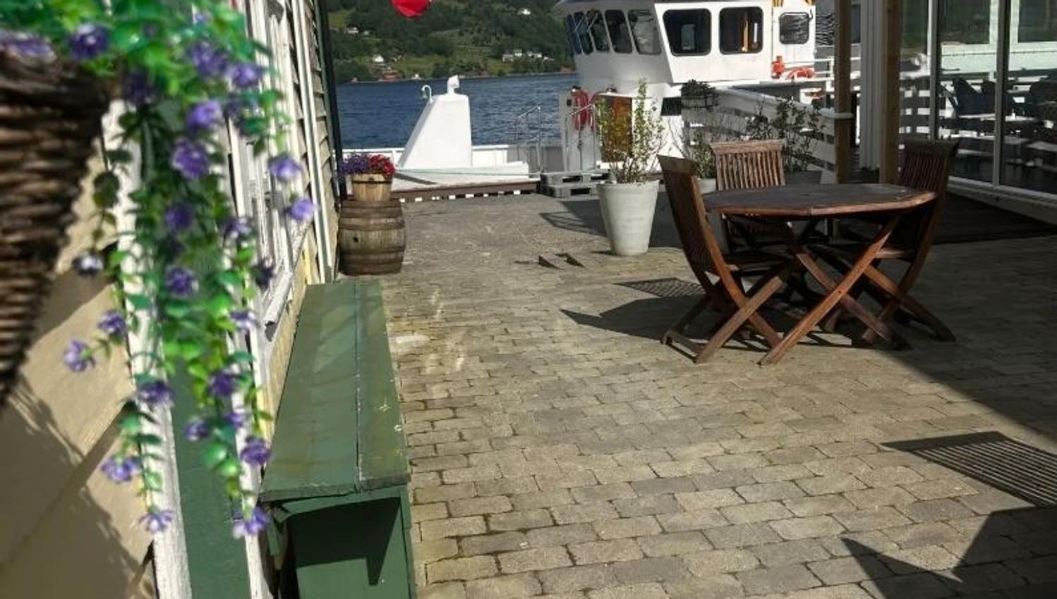 Sunny waterfront patio with wooden table and chairs, potted flowers, a docked boat, and a Norwegian flag..