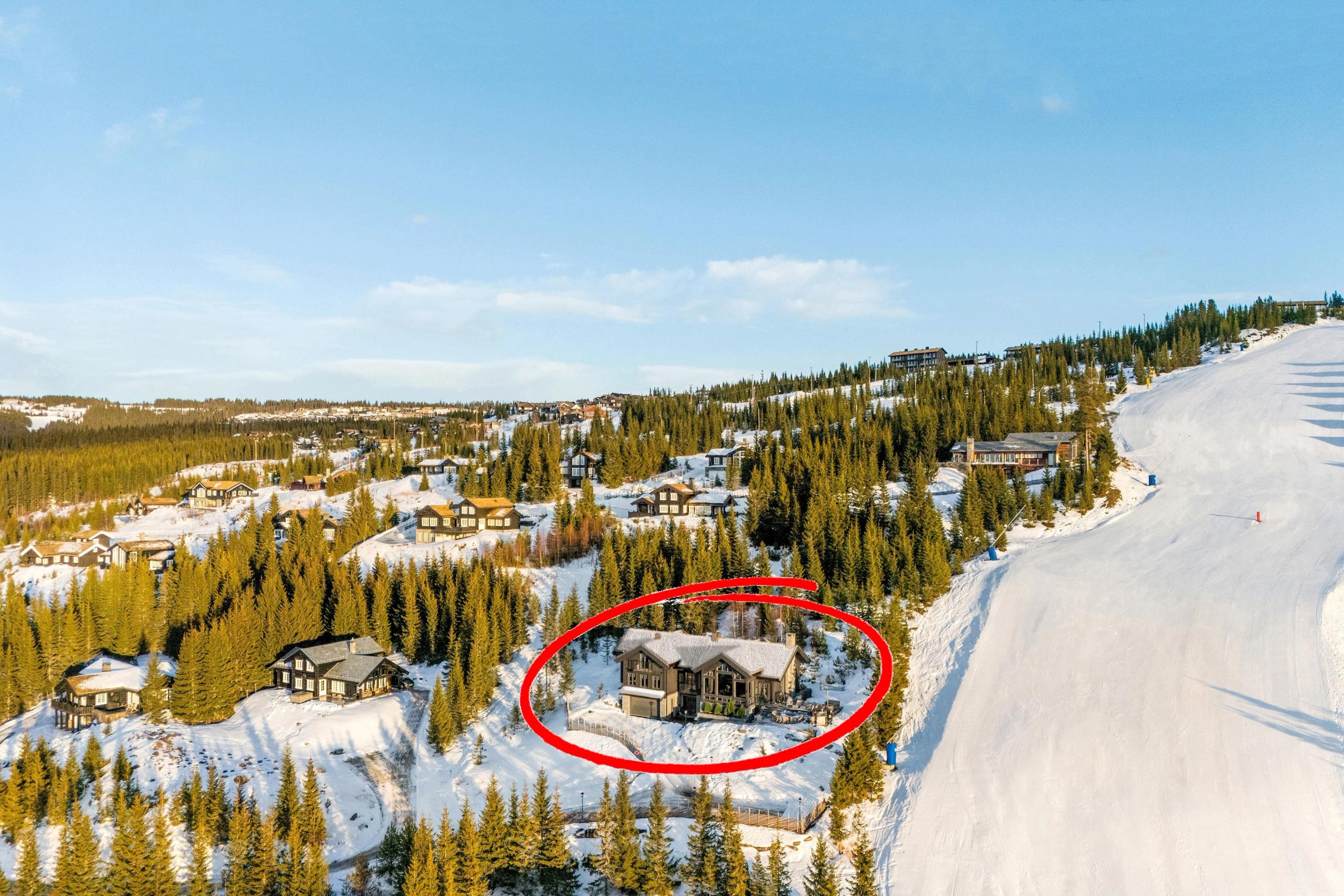Landskap med snødekte hytter i skogkledd fjellside, og en bred skibakke til høyre under blå himmel..