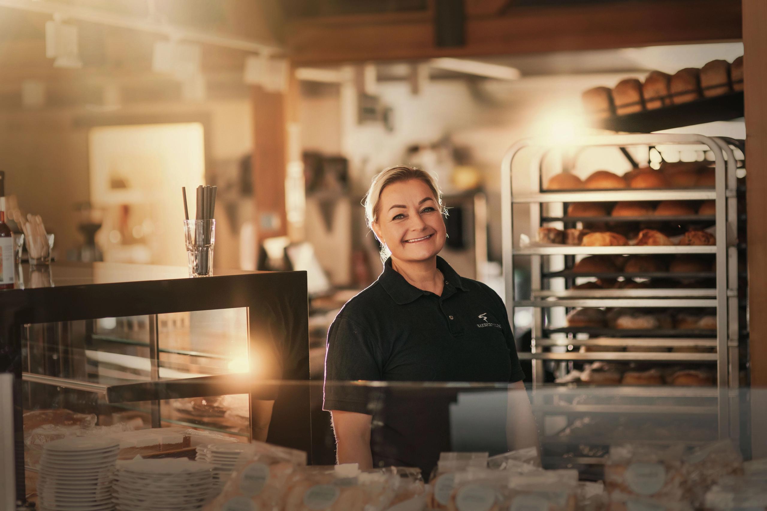A person in a dark polo stands behind a bakery counter, with pastry cases and bread racks in warm light..