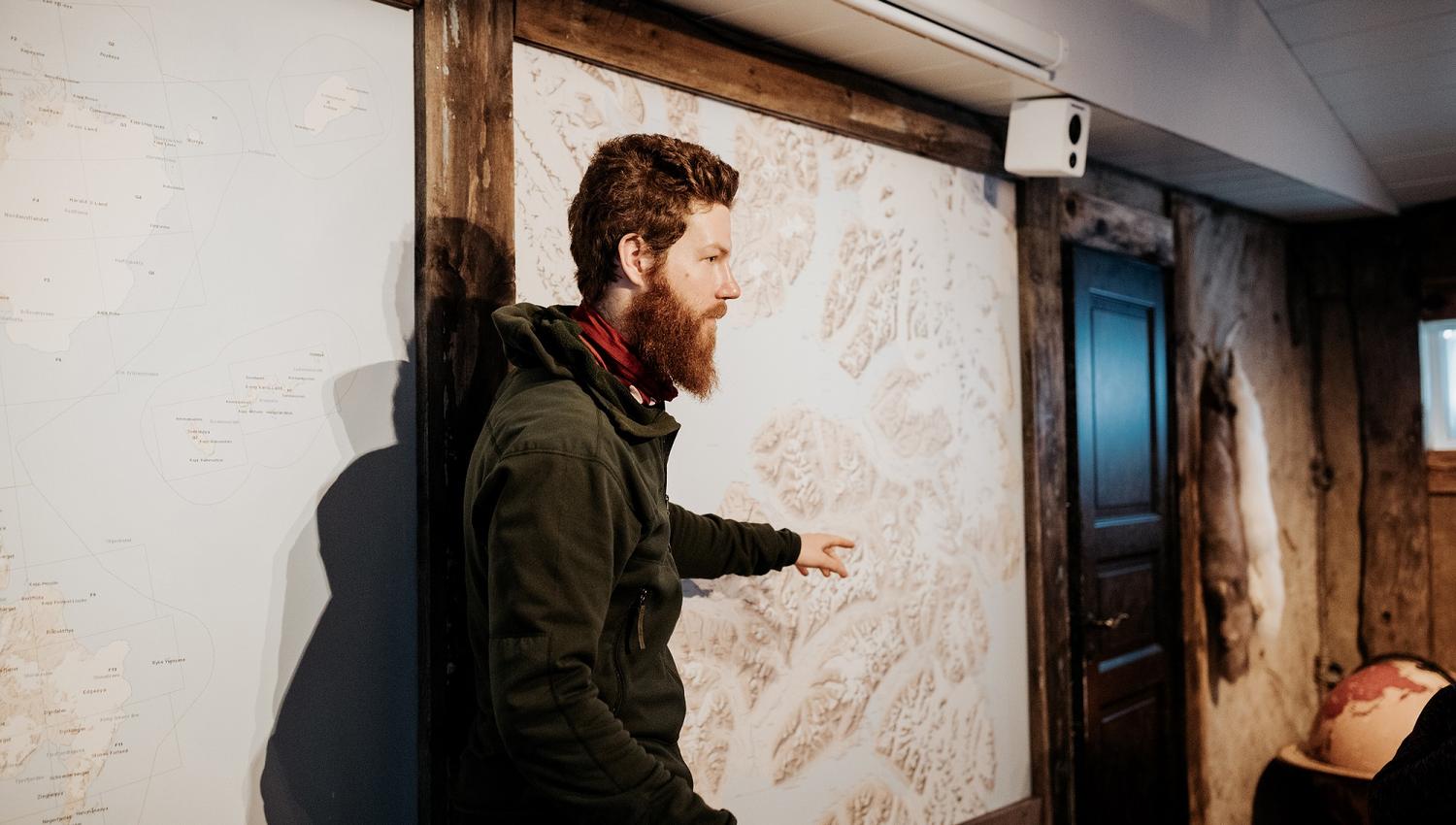 A guide pointing out a trip route on a map hanging on a wall in the background