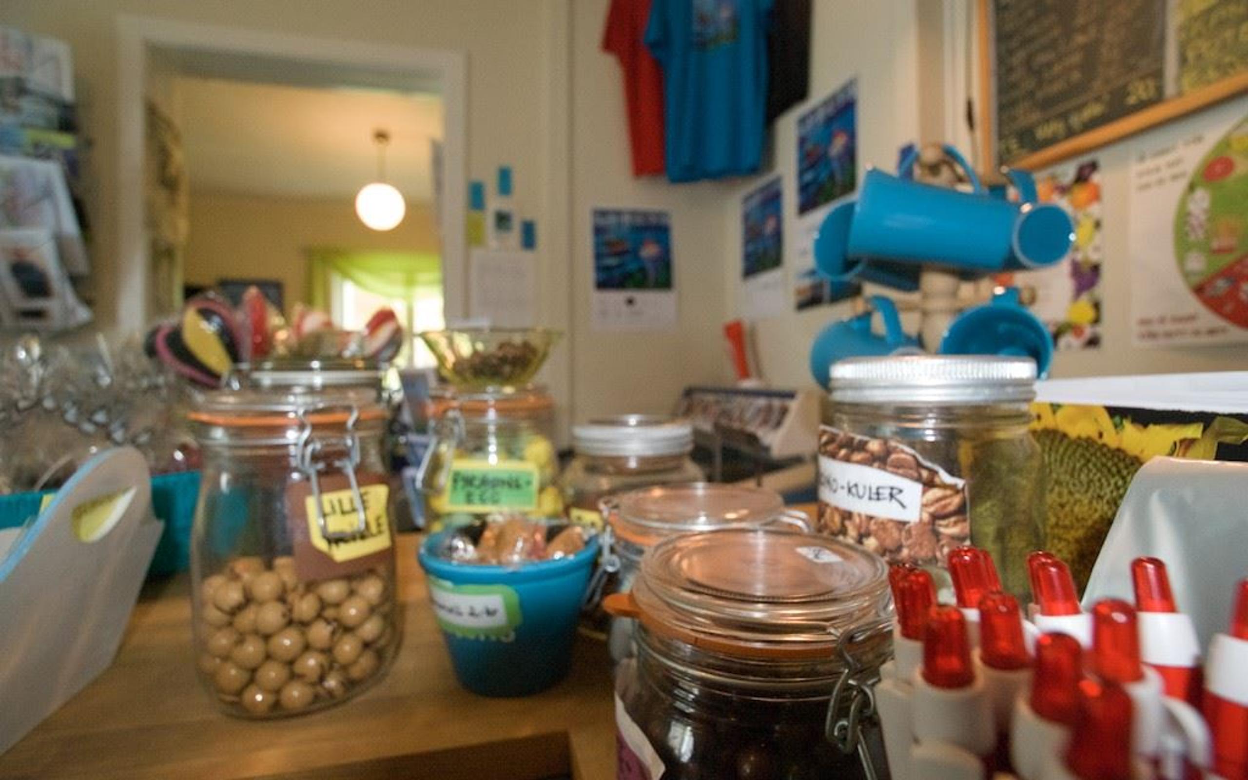 A cluttered shop counter with glass jars of snacks, small tubs, red-capped pens, and posters on the wall behind.