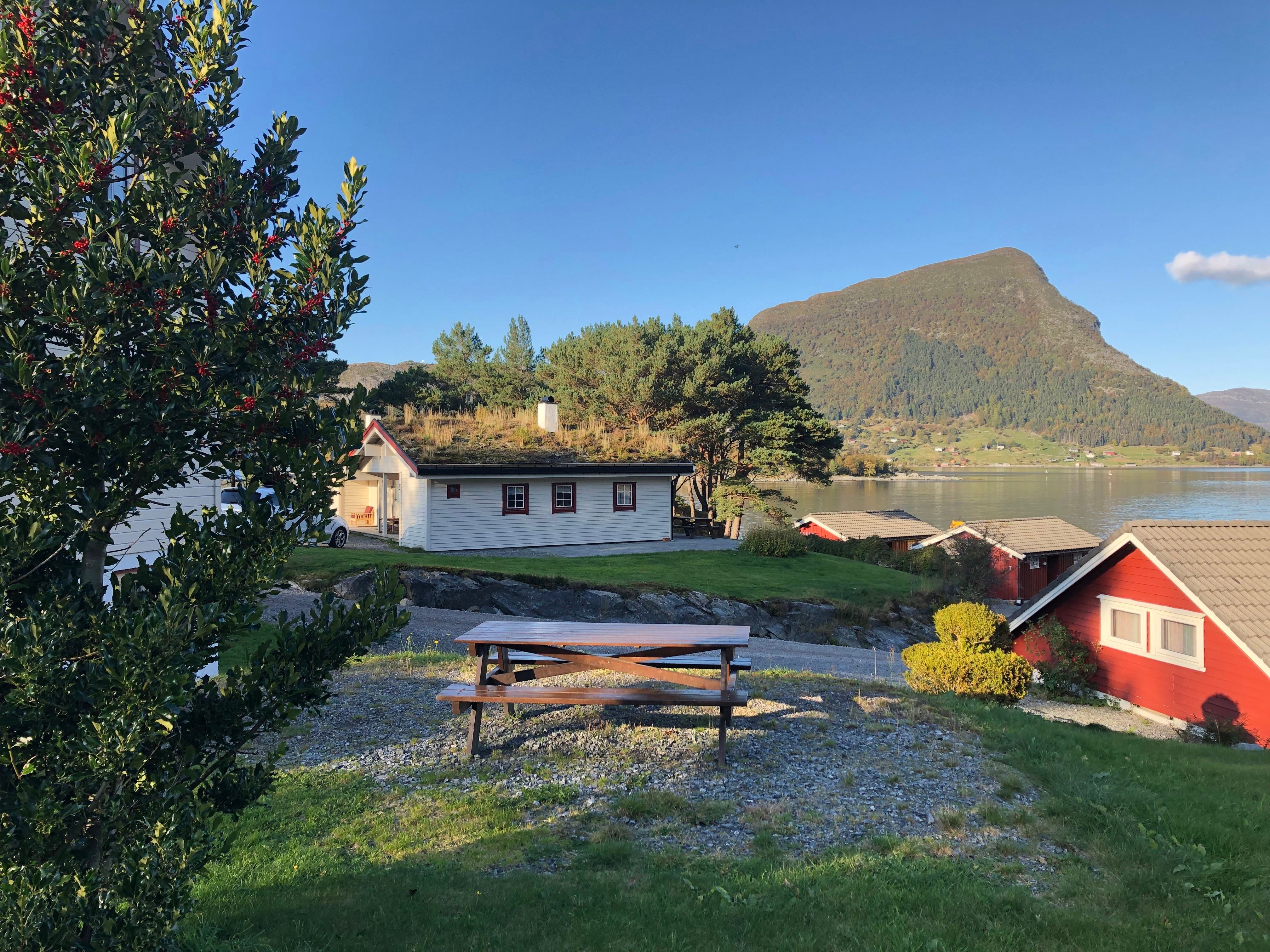 Picnic table on a grassy yard beside red and white cabins, with a lake and forested mountain under a clear sky..