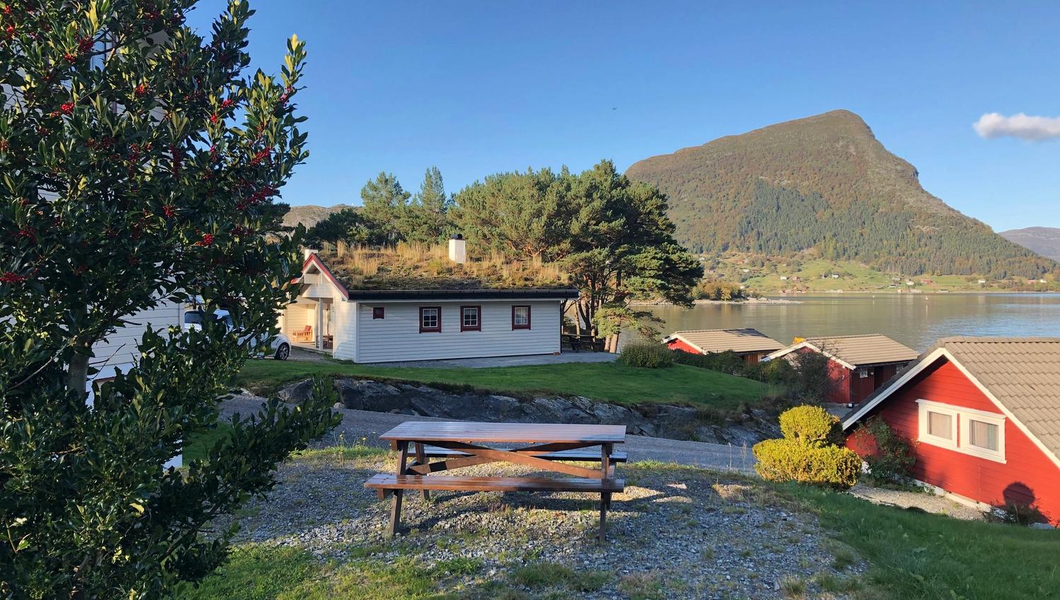 Picnic table on a grassy yard beside red and white cabins, with a lake and forested mountain under a clear sky..