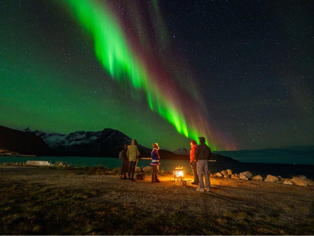Gjester samler seg rundt bålet og ser opp mot nordlyset