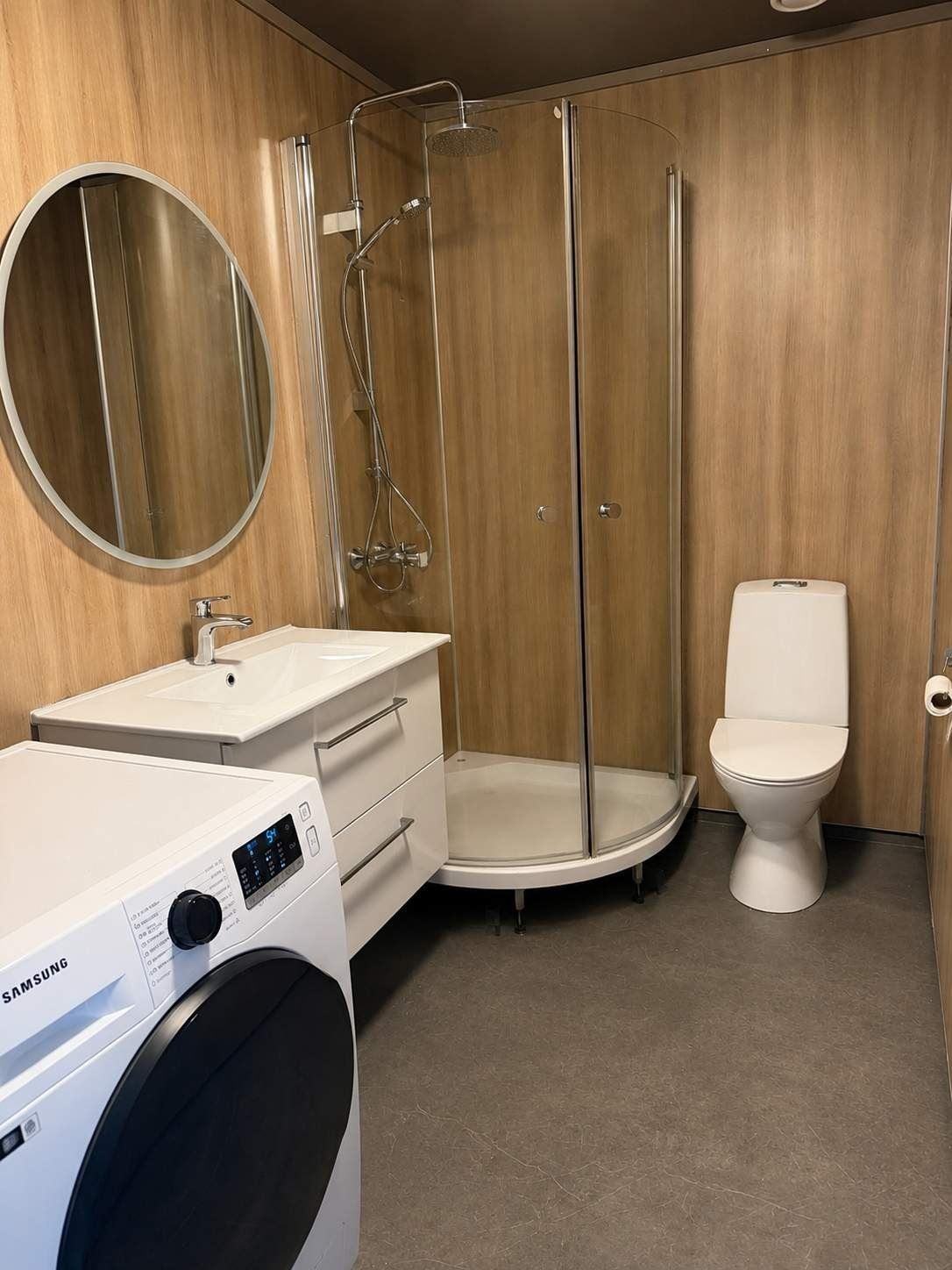 Modern bathroom with wood-paneled walls, round mirror, sink vanity, front-load washer, shower stall, and toilet.