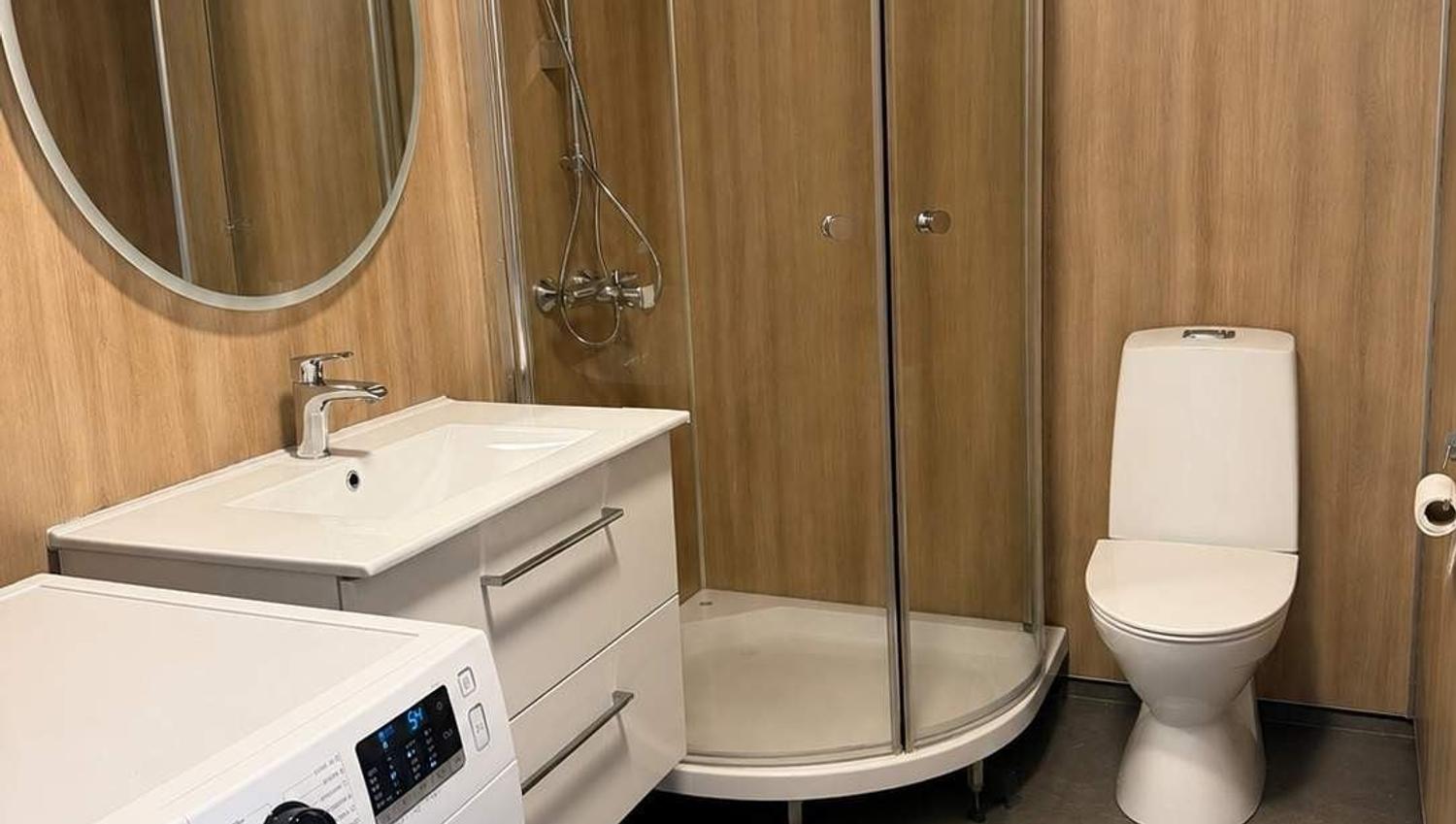 Modern bathroom with wood-paneled walls, round mirror, sink vanity, front-load washer, shower stall, and toilet.