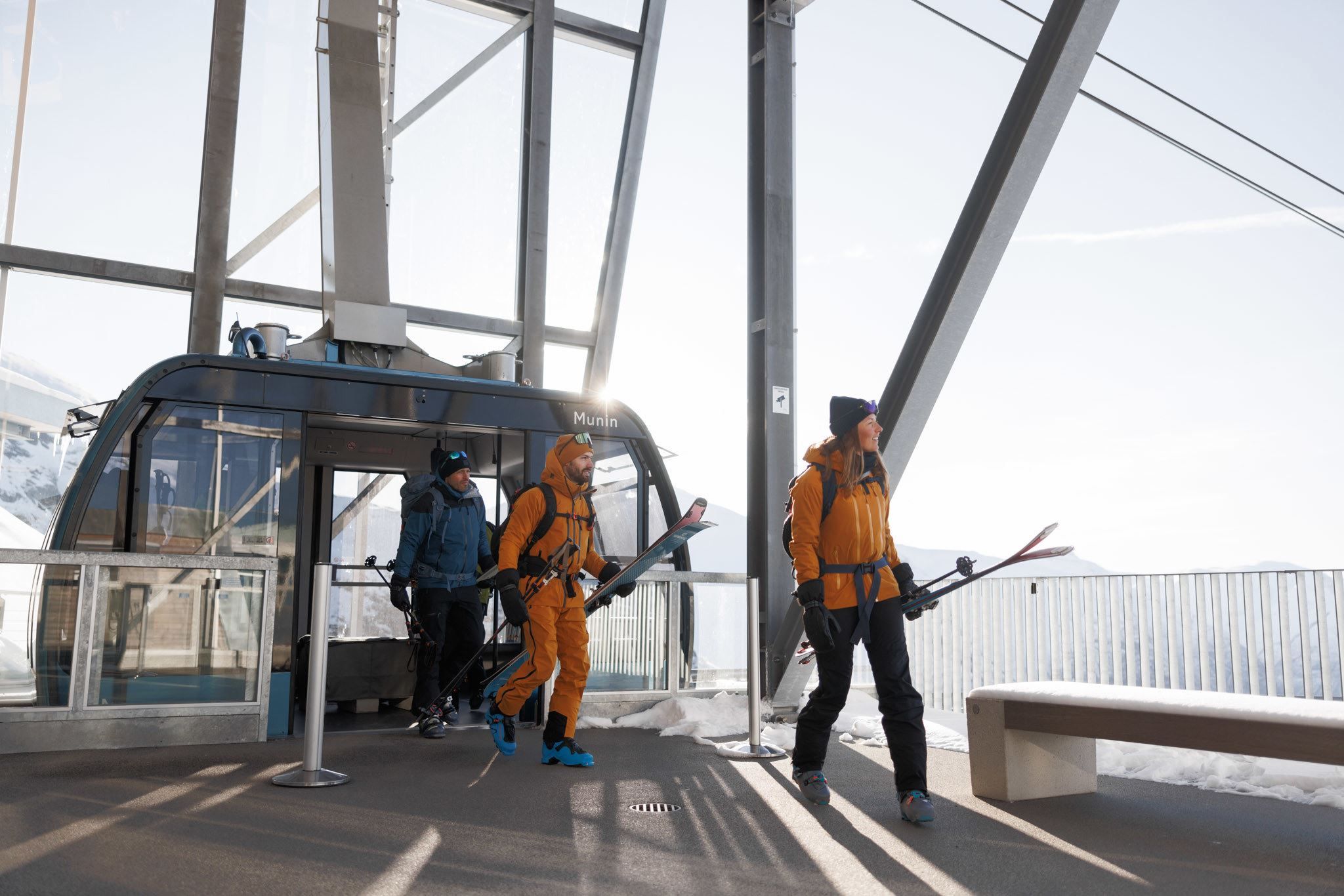 Three skiers in winter gear step out of a gondola station, carrying skis with snow and bright sky outside.
