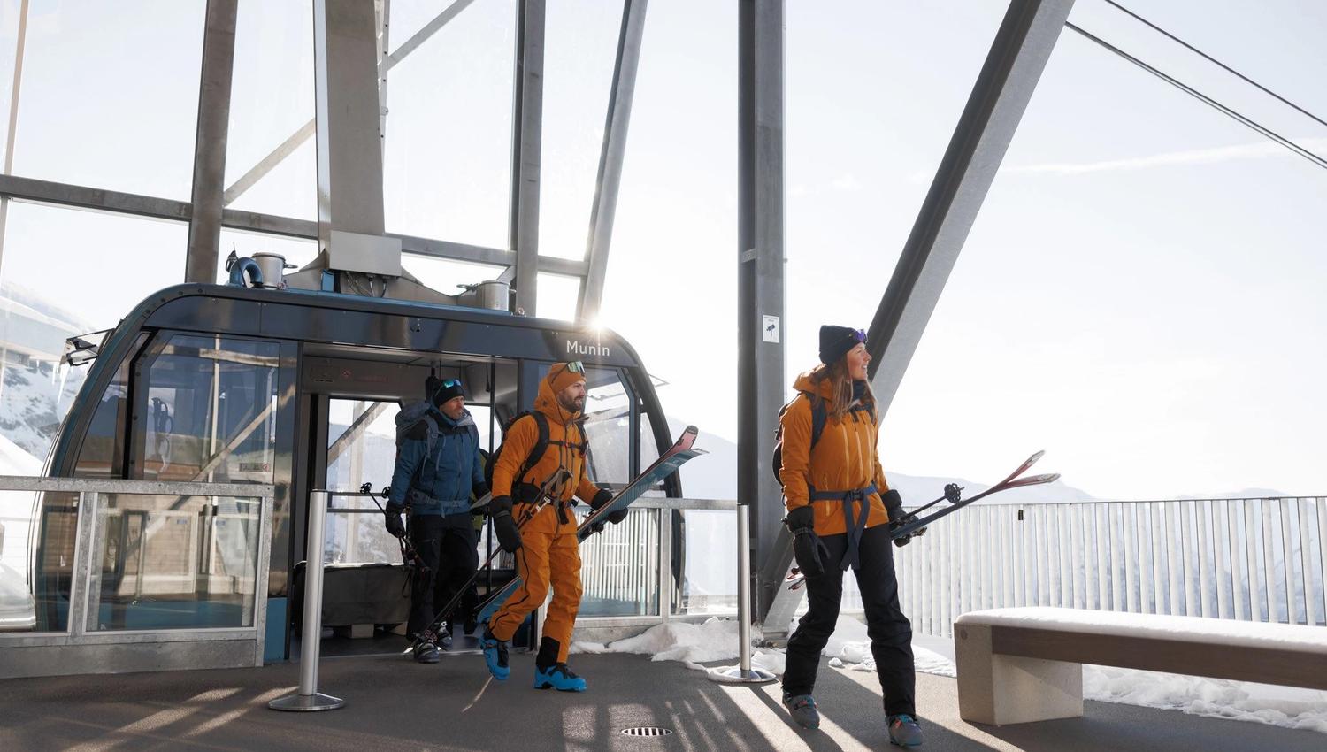 Three skiers in winter gear step out of a gondola station, carrying skis with snow and bright sky outside.