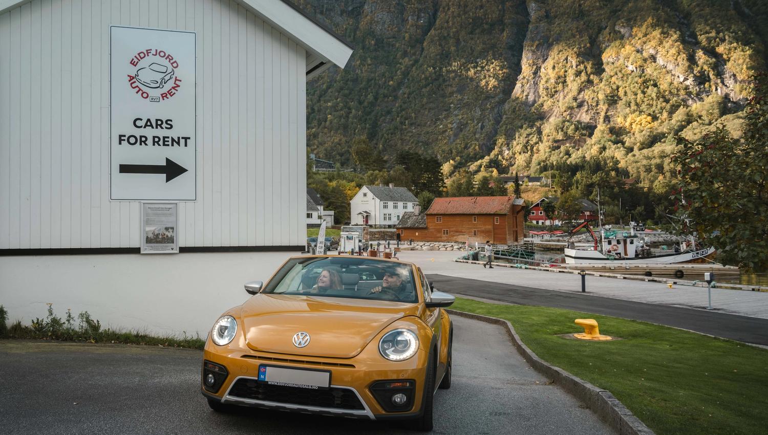 A couple in a gold colored VW beetle car underneath a sign that says Cars for rent.