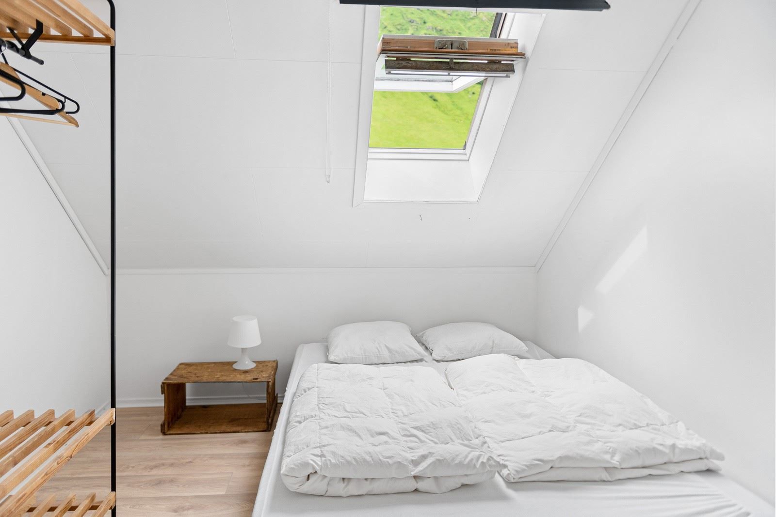 Minimal white attic bedroom with a double bed, wooden nightstand and lamp, open closet rack, and skylight window.