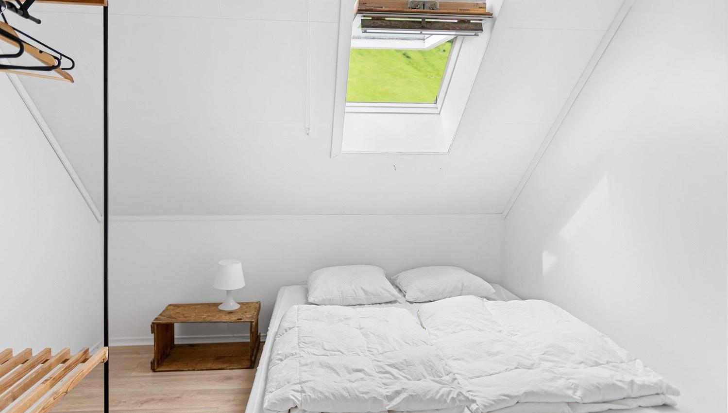 Minimal white attic bedroom with a double bed, wooden nightstand and lamp, open closet rack, and skylight window.