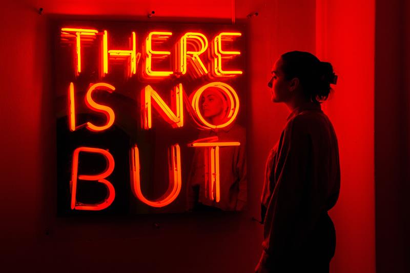 Person standing in front of a red neon sign in a darkened exhibition space.