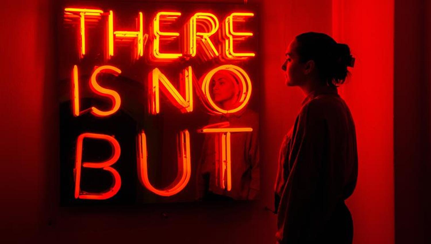 Person standing in front of a red neon sign in a darkened exhibition space.