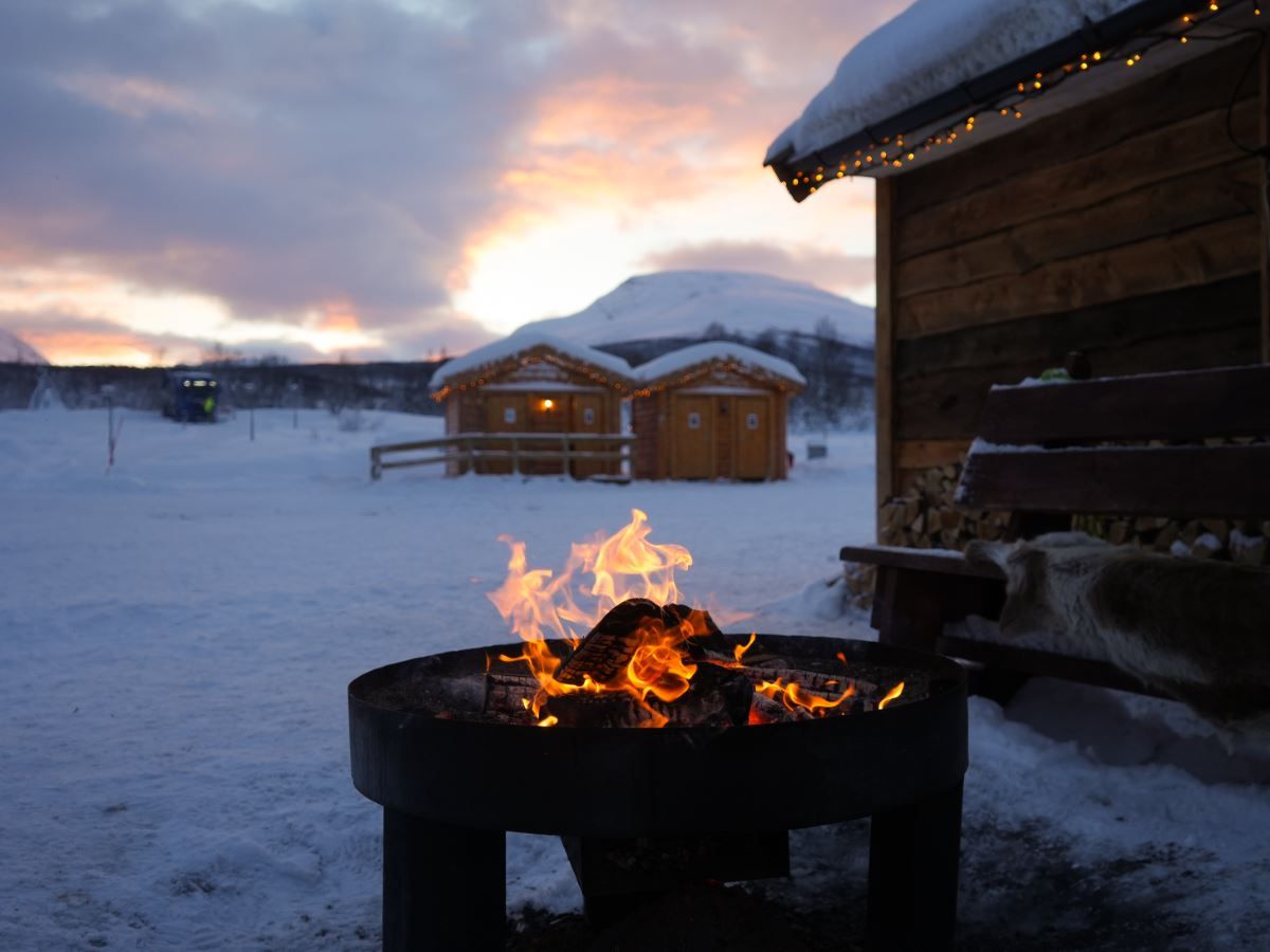 Outdoor fire at Tromsø Lapland’s Sámi camp with snow-covered buildings and winter scenery in the background.