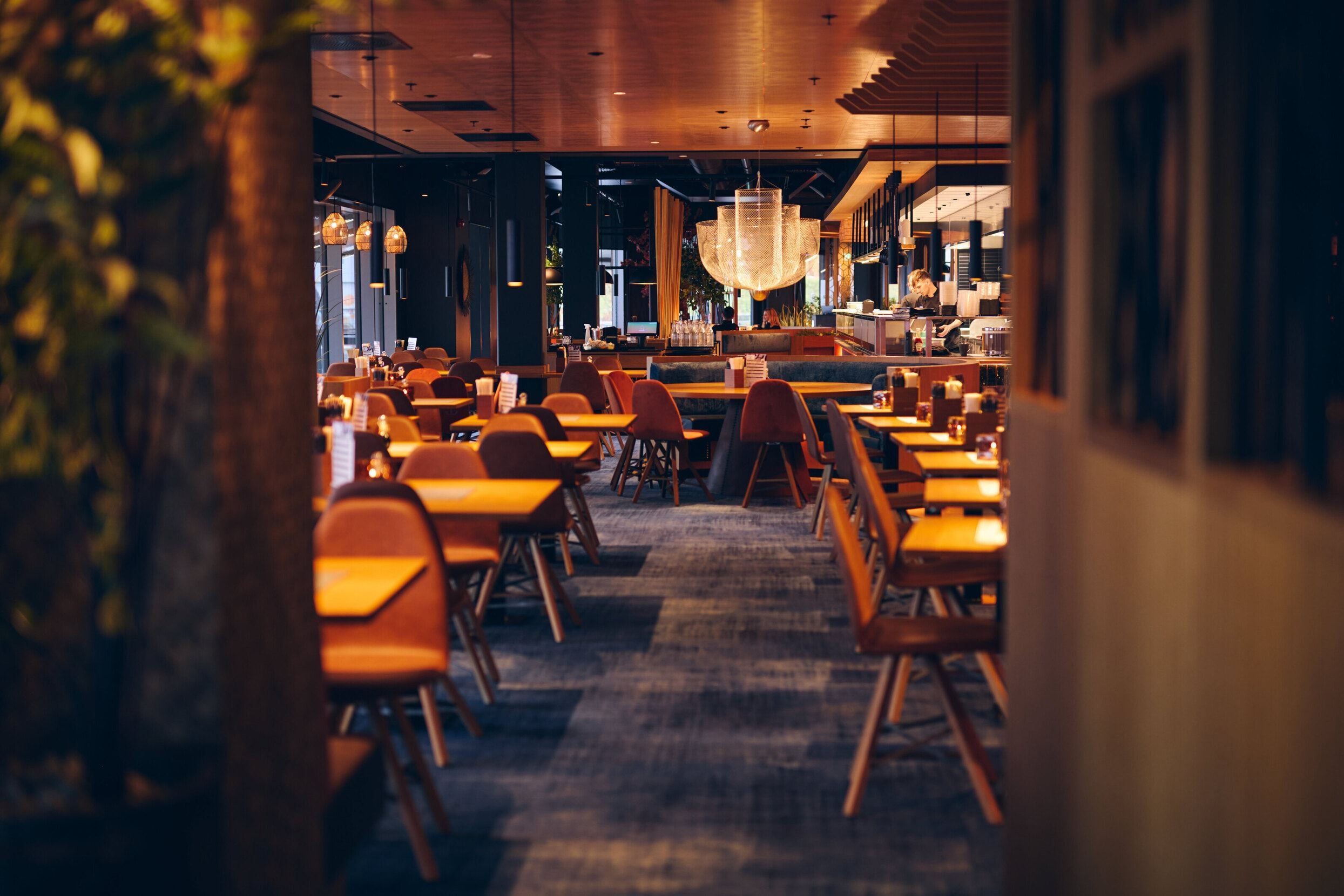 Moderne restaurantinteriør med trebord og stoler, stor lysekrone og varm belysning i et rolig og innbydende miljø.