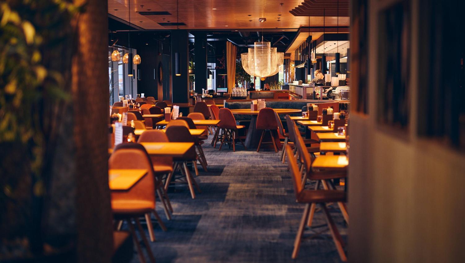 Moderne restaurantinteriør med trebord og stoler, stor lysekrone og varm belysning i et rolig og innbydende miljø.
