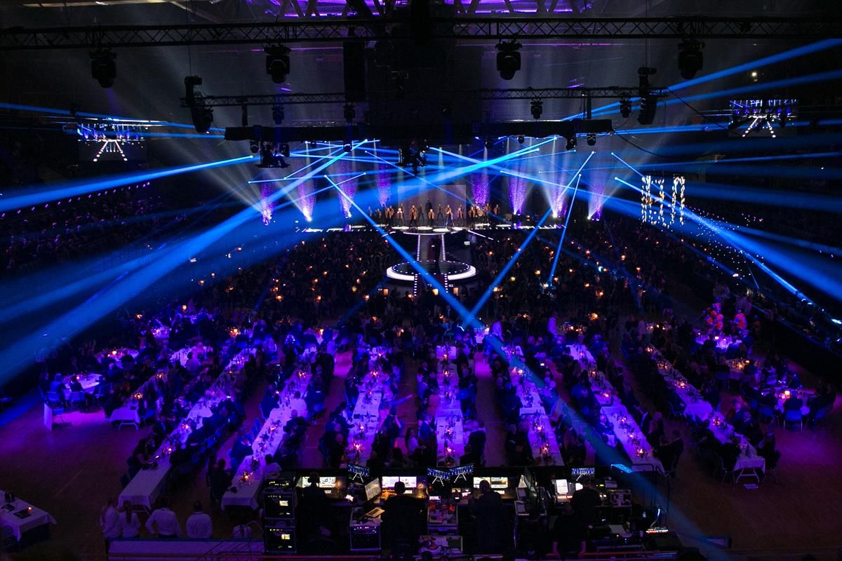 Concert hall filled with an audience, illuminated by blue stage lights and laser beams shining across the room.