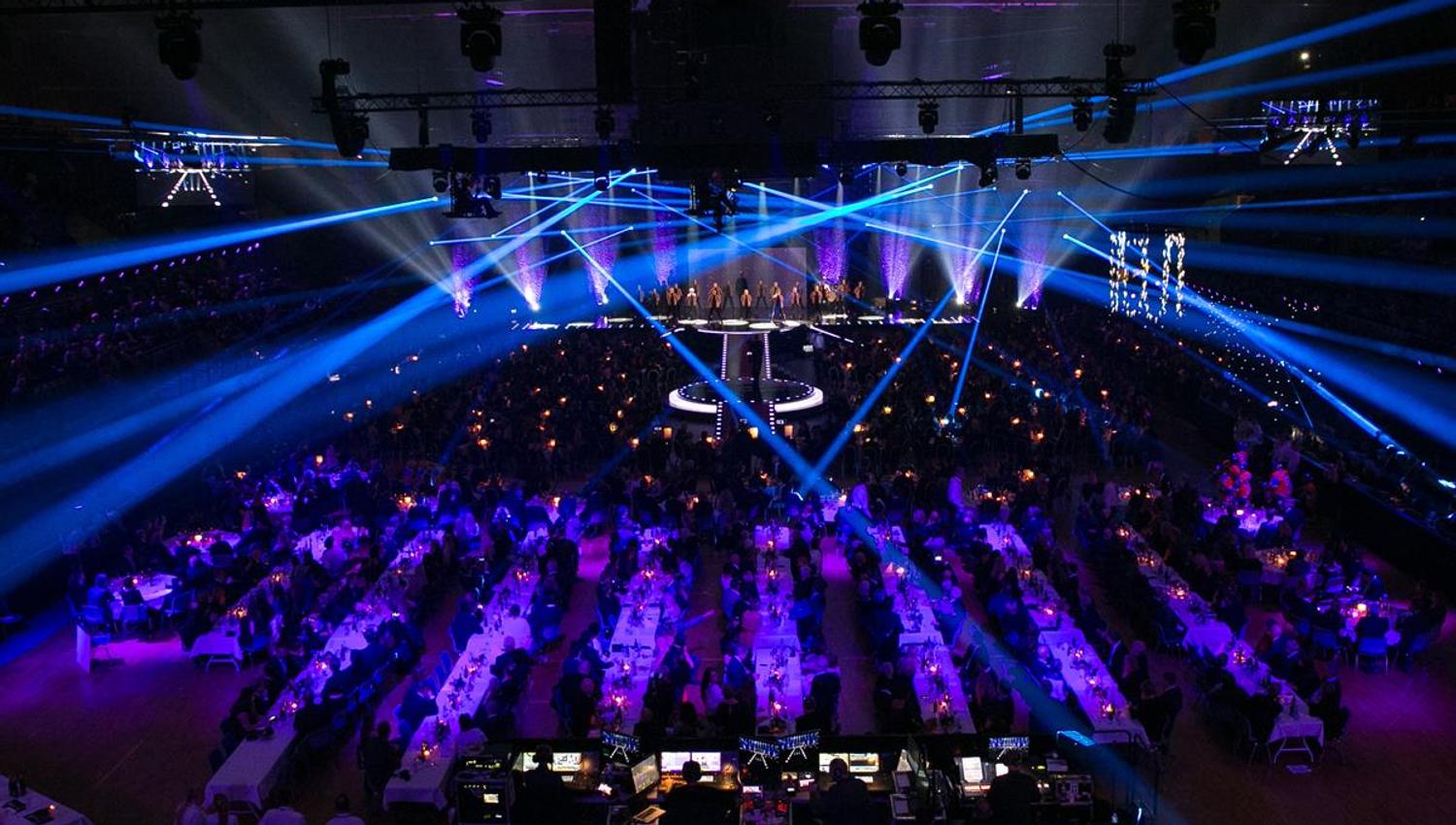 Concert hall filled with an audience, illuminated by blue stage lights and laser beams shining across the room.