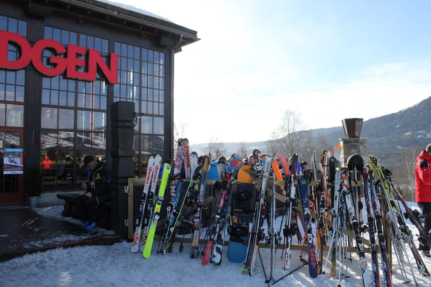Mange ski og snowboard står i stativ utenfor en bygning med store vinduer, med mennesker og fjell i bakgrunnen..