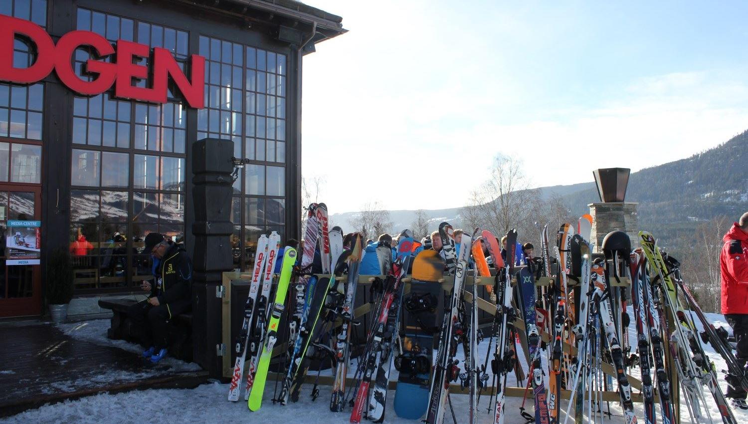 Mange ski og snowboard står i stativ utenfor en bygning med store vinduer, med mennesker og fjell i bakgrunnen..