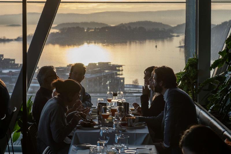 People enjoying a meal at a long table in a restaurant with sunset views over the Oslofjord through large windows