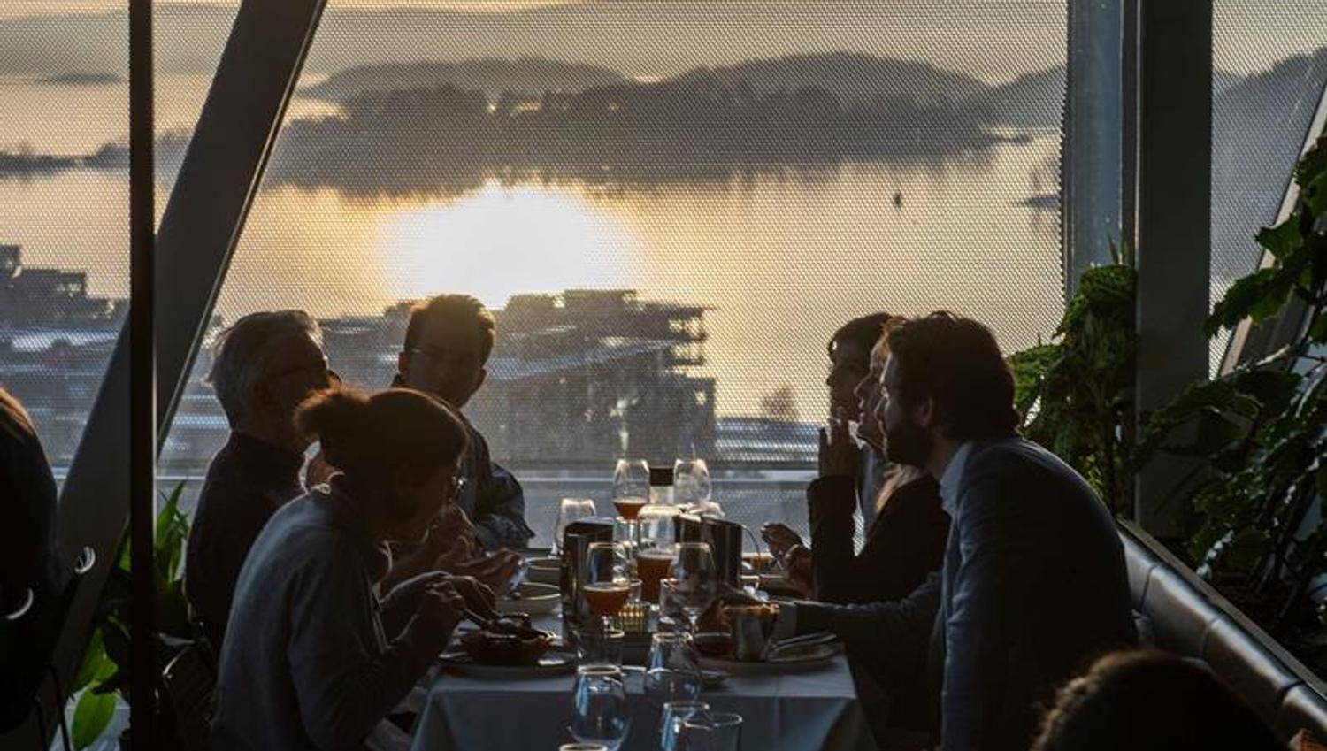 People enjoying a meal at a long table in a restaurant with sunset views over the Oslofjord through large windows