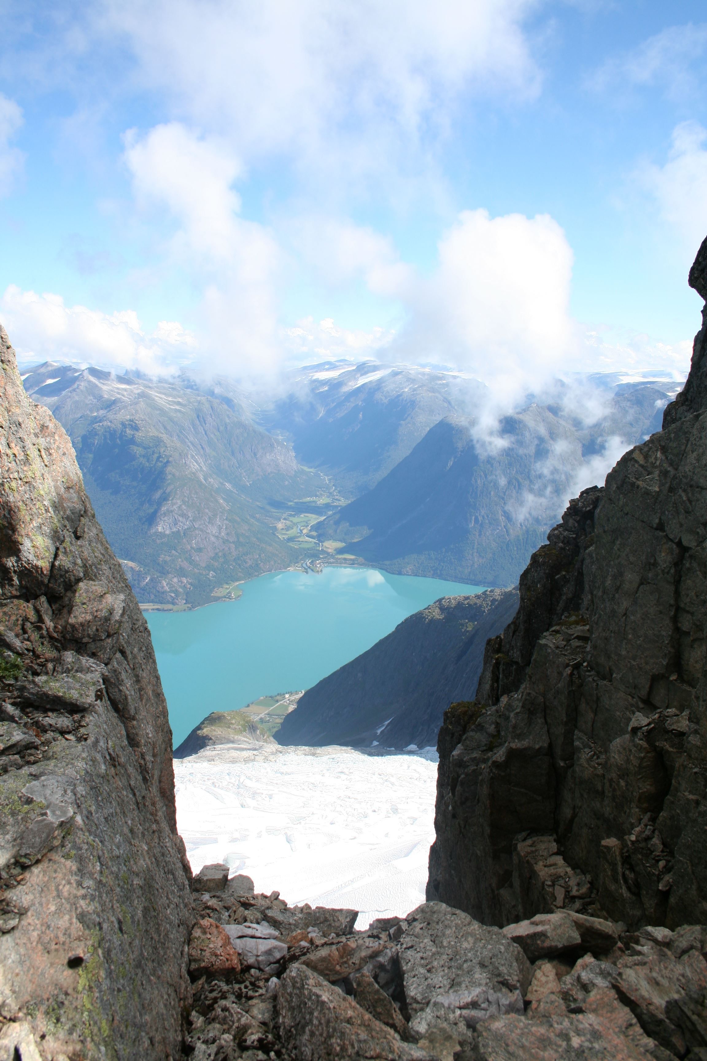 View through rocky cliffs to a turquoise lake below, with distant mountains, snowfields, and scattered clouds.
