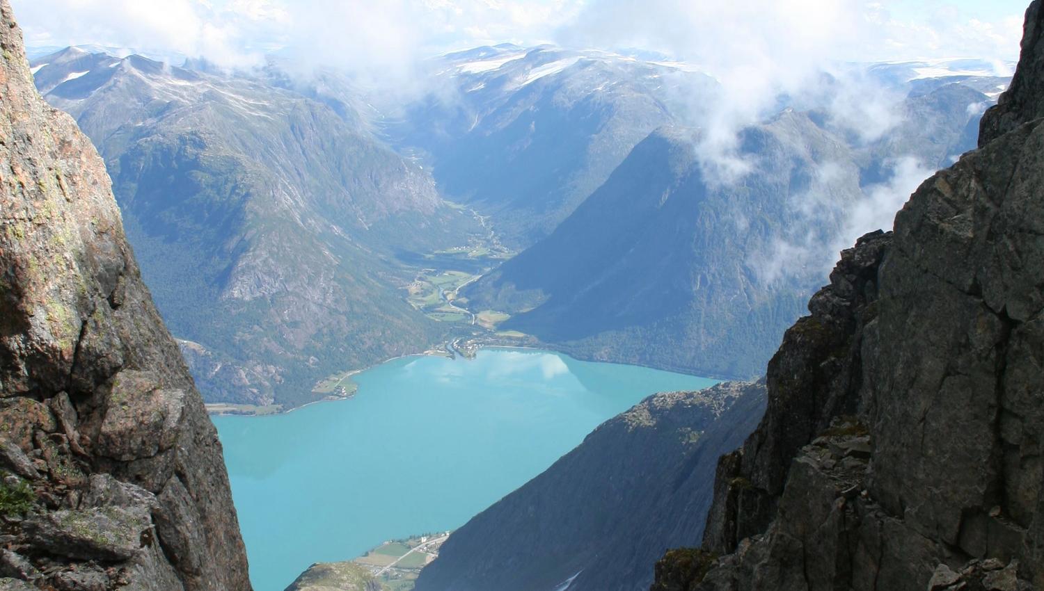 View through rocky cliffs to a turquoise lake below, with distant mountains, snowfields, and scattered clouds.