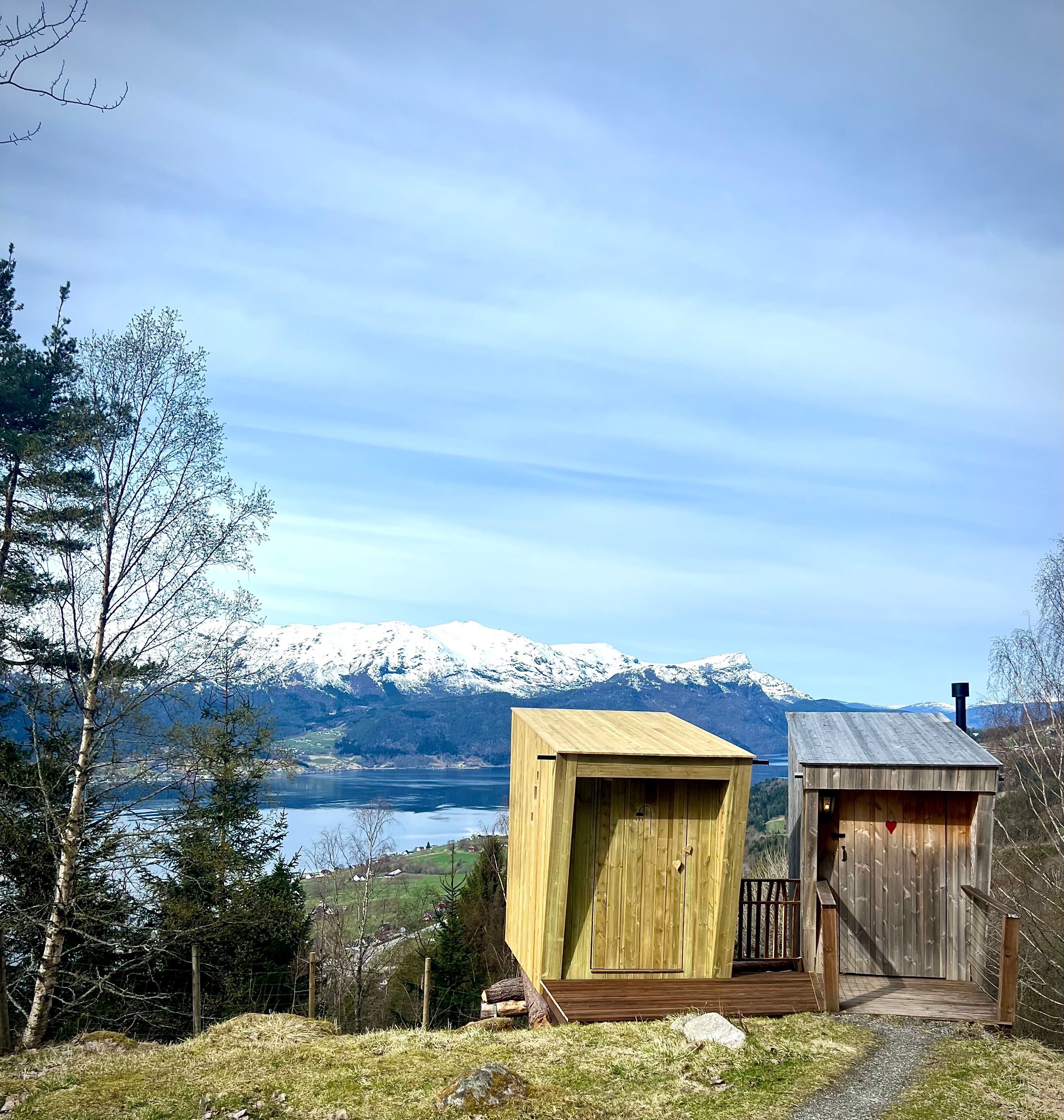 Two small wooden cabins on a grassy hillside overlook a lake and snow-capped mountains under a cloudy sky..