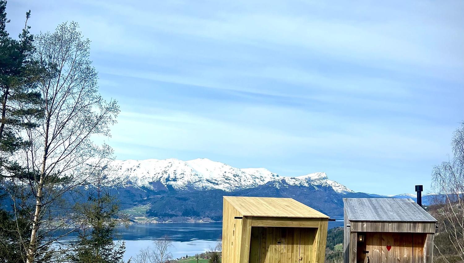 Two small wooden cabins on a grassy hillside overlook a lake and snow-capped mountains under a cloudy sky..