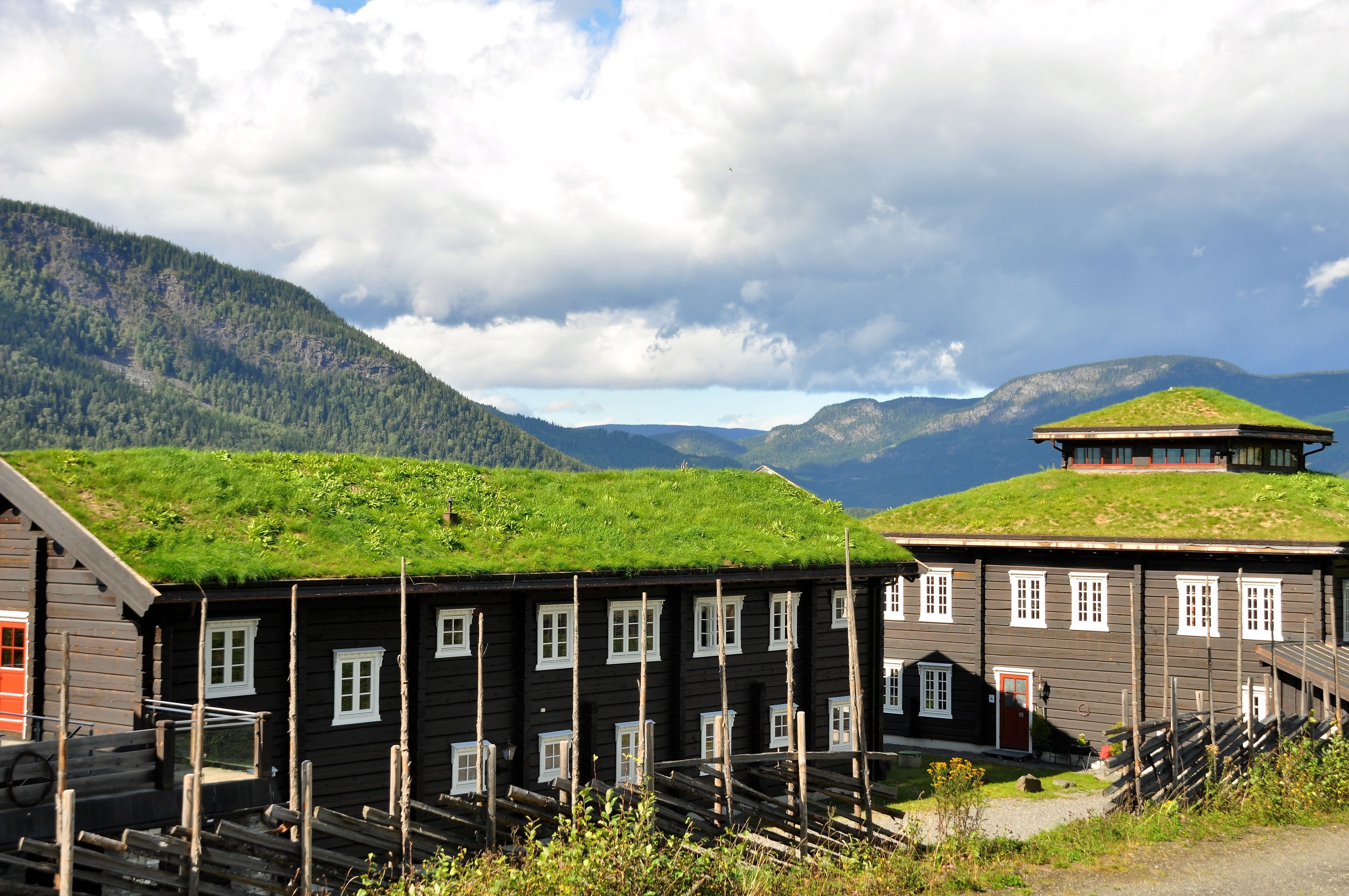 Mørke trehus med hvitmalte vinduer og torvtak i sollys, med fjell og store skyer i bakgrunnen..