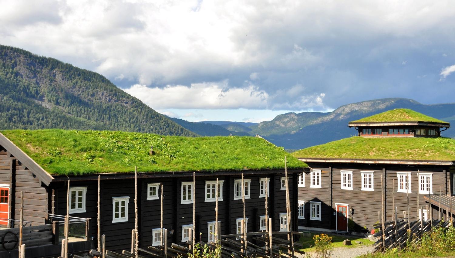Mørke trehus med hvitmalte vinduer og torvtak i sollys, med fjell og store skyer i bakgrunnen..