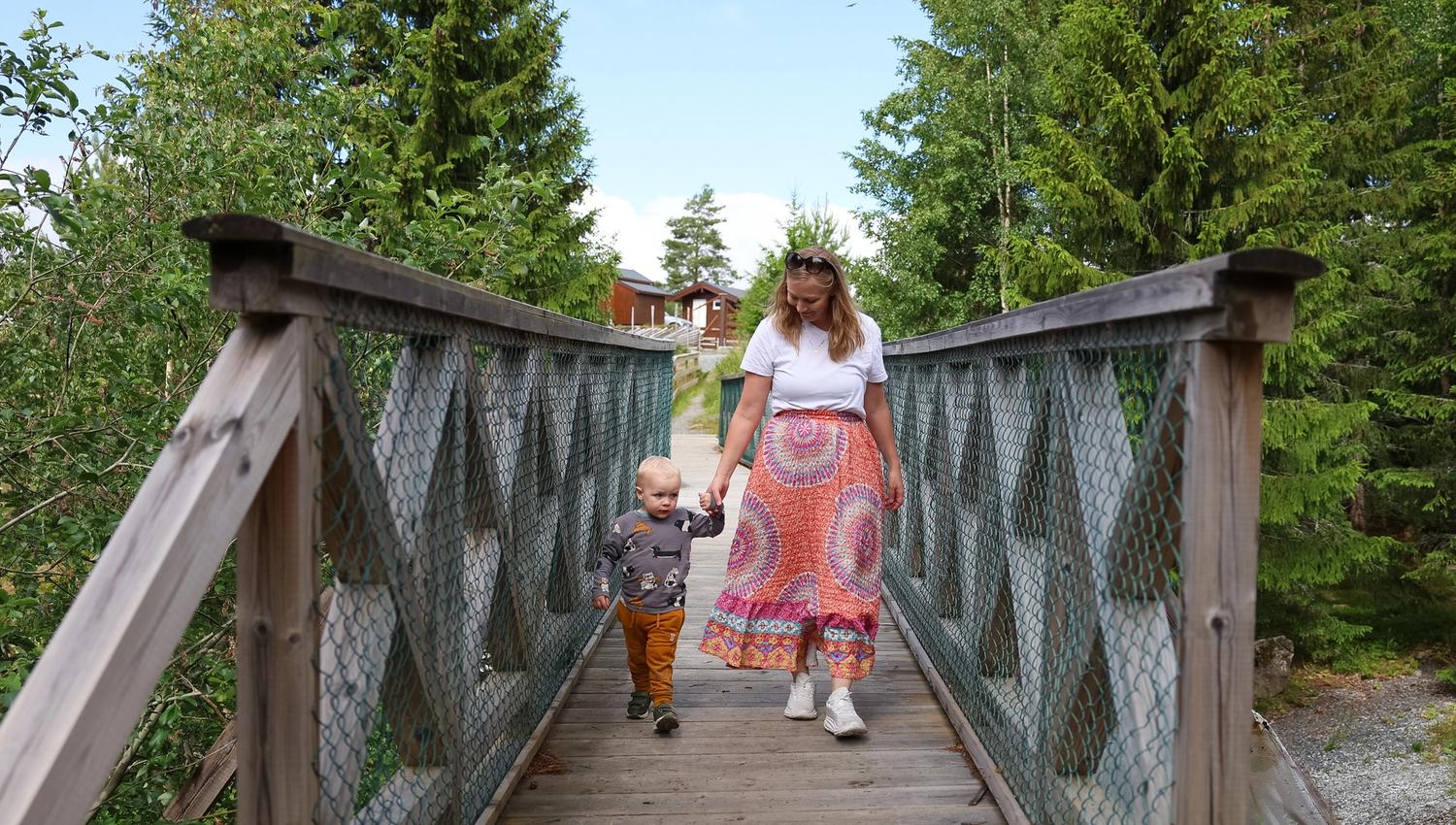 Barn og mamma som går over en trebro omgitt av grønne trær i naturparken