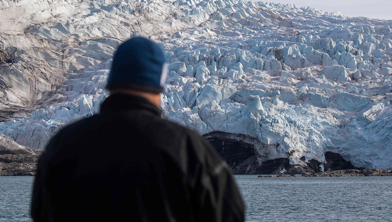A guest standing on a boat in the foreground looking out at a glacier in the background