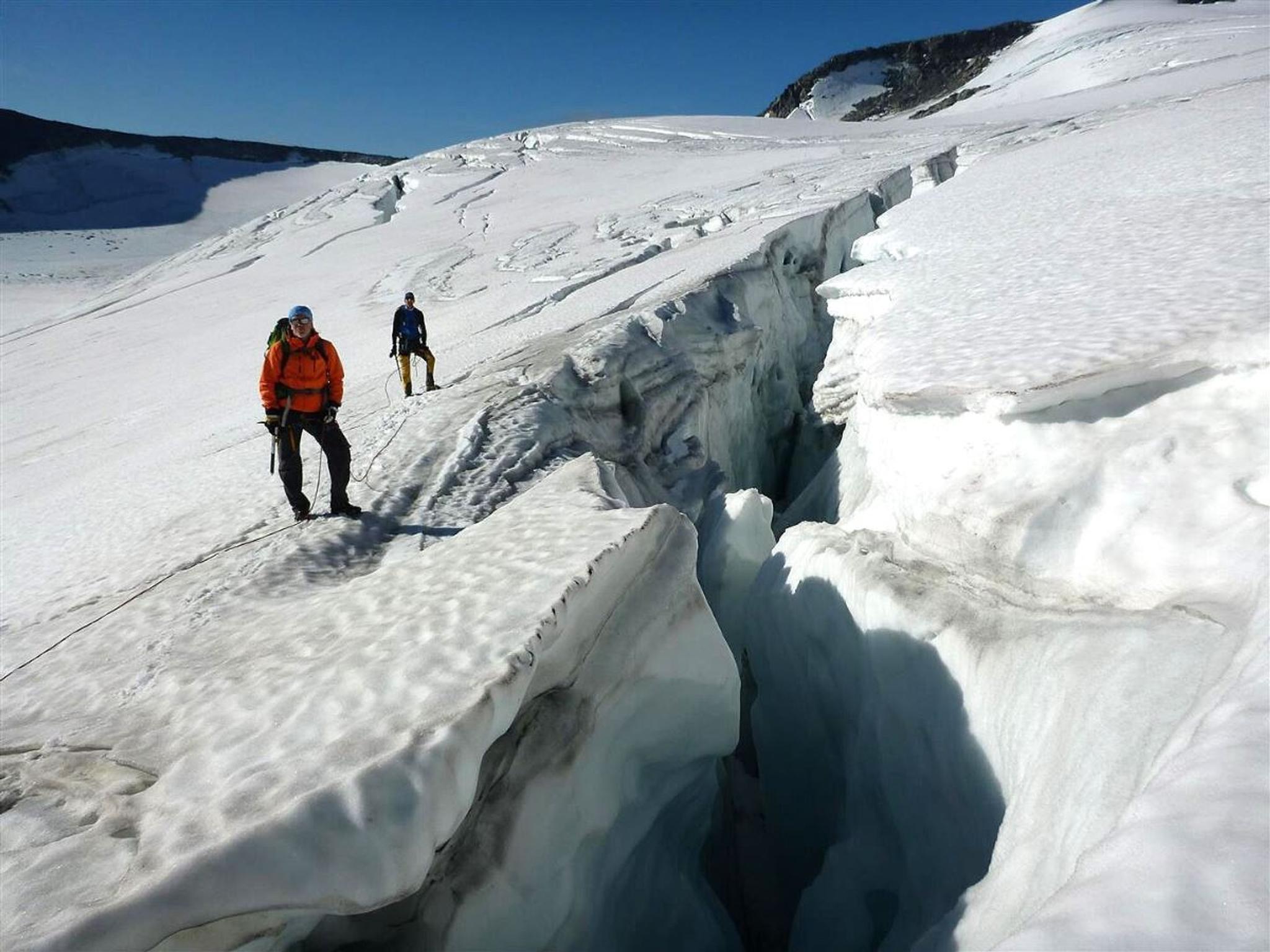 Two glacier walkers with ropes and ice axes stand on a snow-covered glacier at a deep crevasse under clear blue skies.