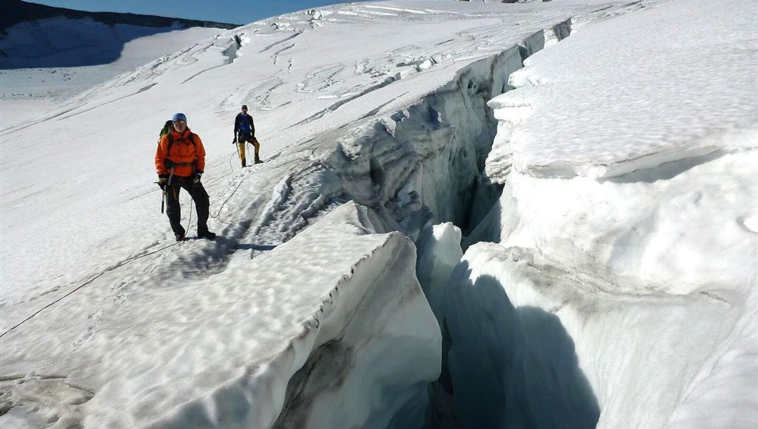 Two glacier walkers with ropes and ice axes stand on a snow-covered glacier at a deep crevasse under clear blue skies.