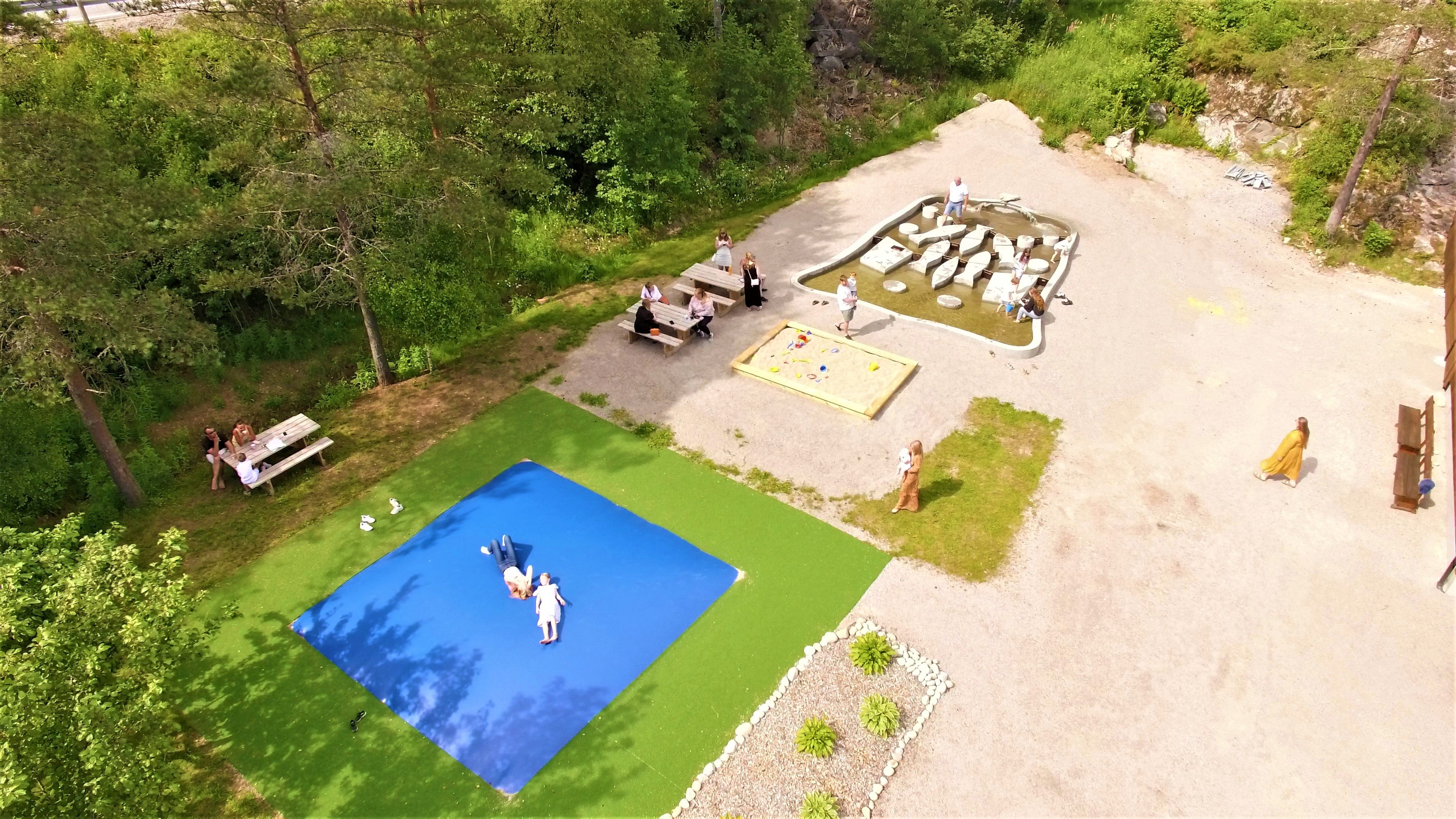 Übersicht über den Spielplatz vor dem Wildlachs-Besucherzentrum in Laudal, mit einem Hüpfkissen, Sandkasten und einem kleinen Teich, über den man auf lachsförmigen Elementen springen kann.