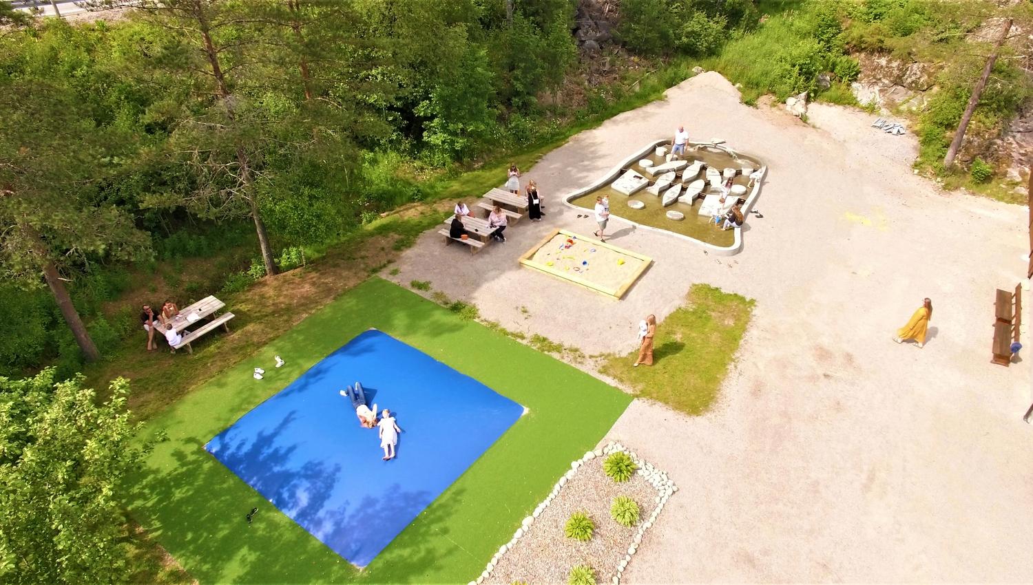 Übersicht über den Spielplatz vor dem Wildlachs-Besucherzentrum in Laudal, mit einem Hüpfkissen, Sandkasten und einem kleinen Teich, über den man auf lachsförmigen Elementen springen kann.