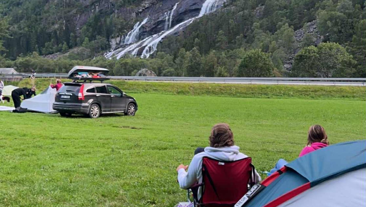 Gäste entspannen sich am Zelt auf dem Hildal Camping mit dem Wasserfall als Kulisse.