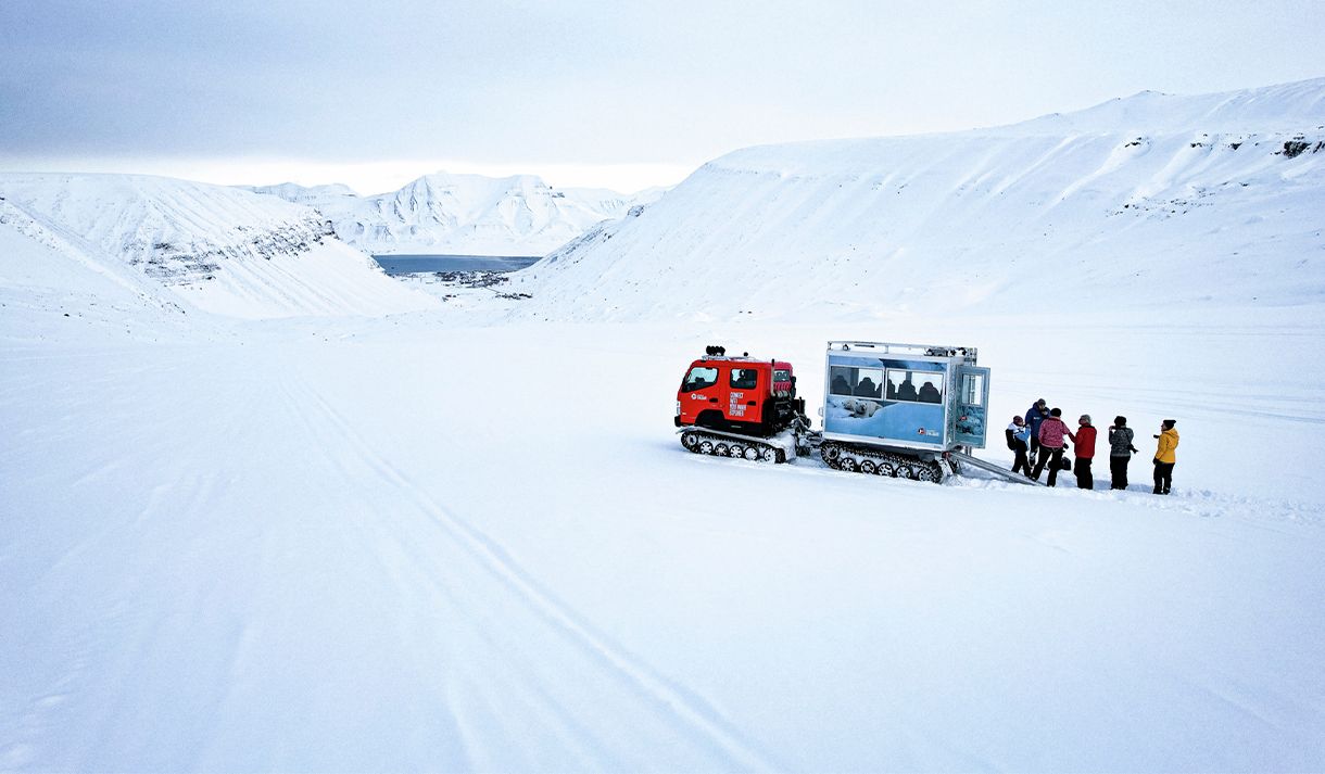 Gjester og en guide som står ved siden av en beltevogn på en isbre omringet av et snødekt fjellandskap