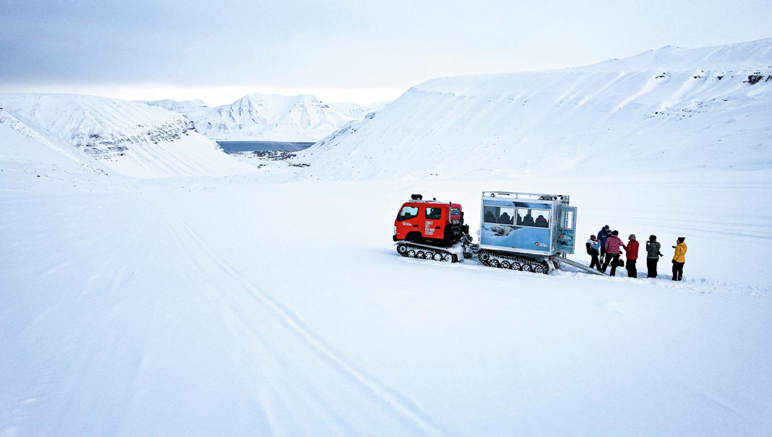 Gjester og en guide som står ved siden av en beltevogn på en isbre omringet av et snødekt fjellandskap