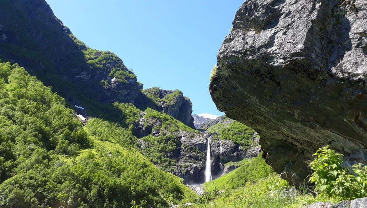 A narrow waterfall drops between rocky cliffs in a lush green valley, with a large rock overhang at right under a blue sky..