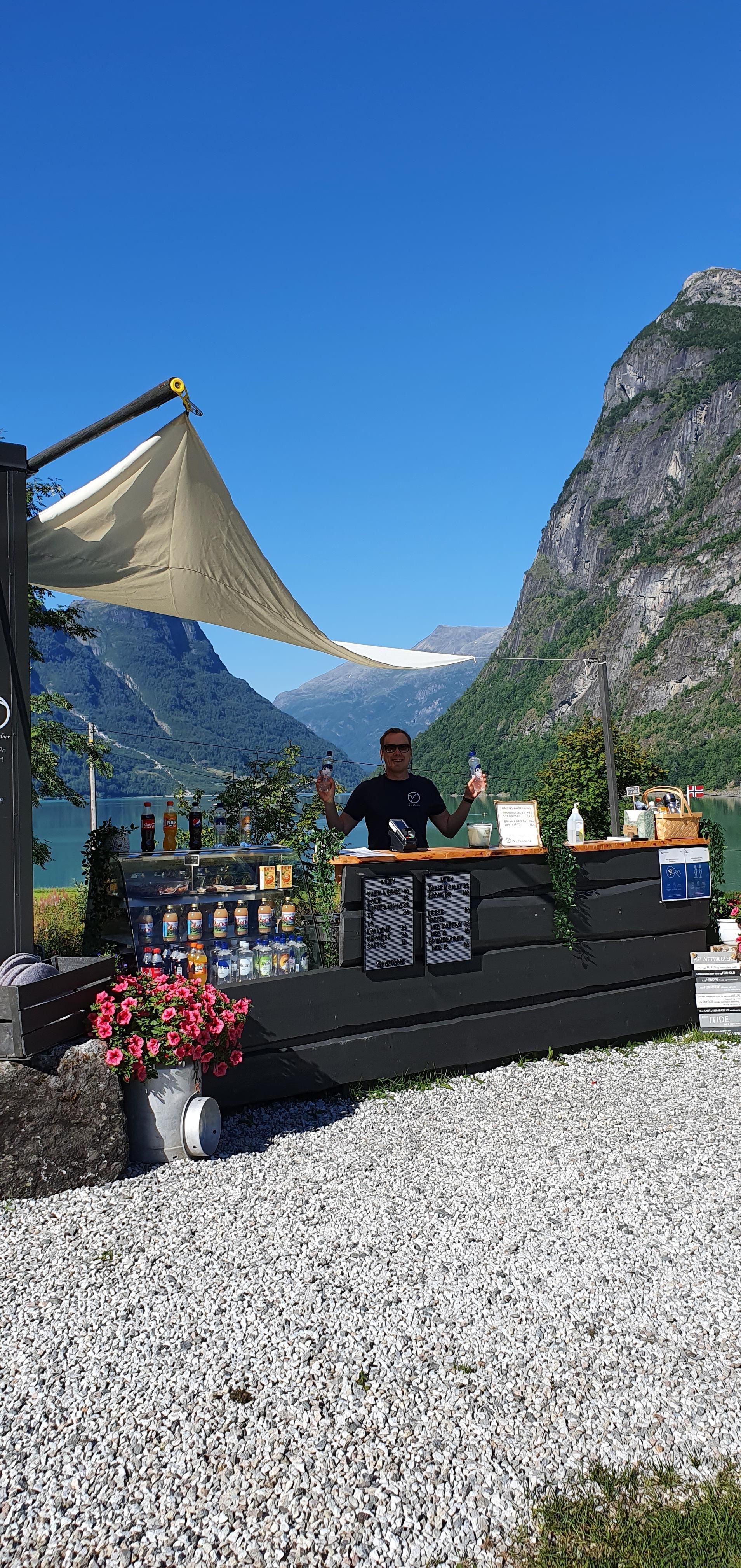 Outdoor drink kiosk with a person behind the counter, bottles and flowers, set by a lake with steep mountains.